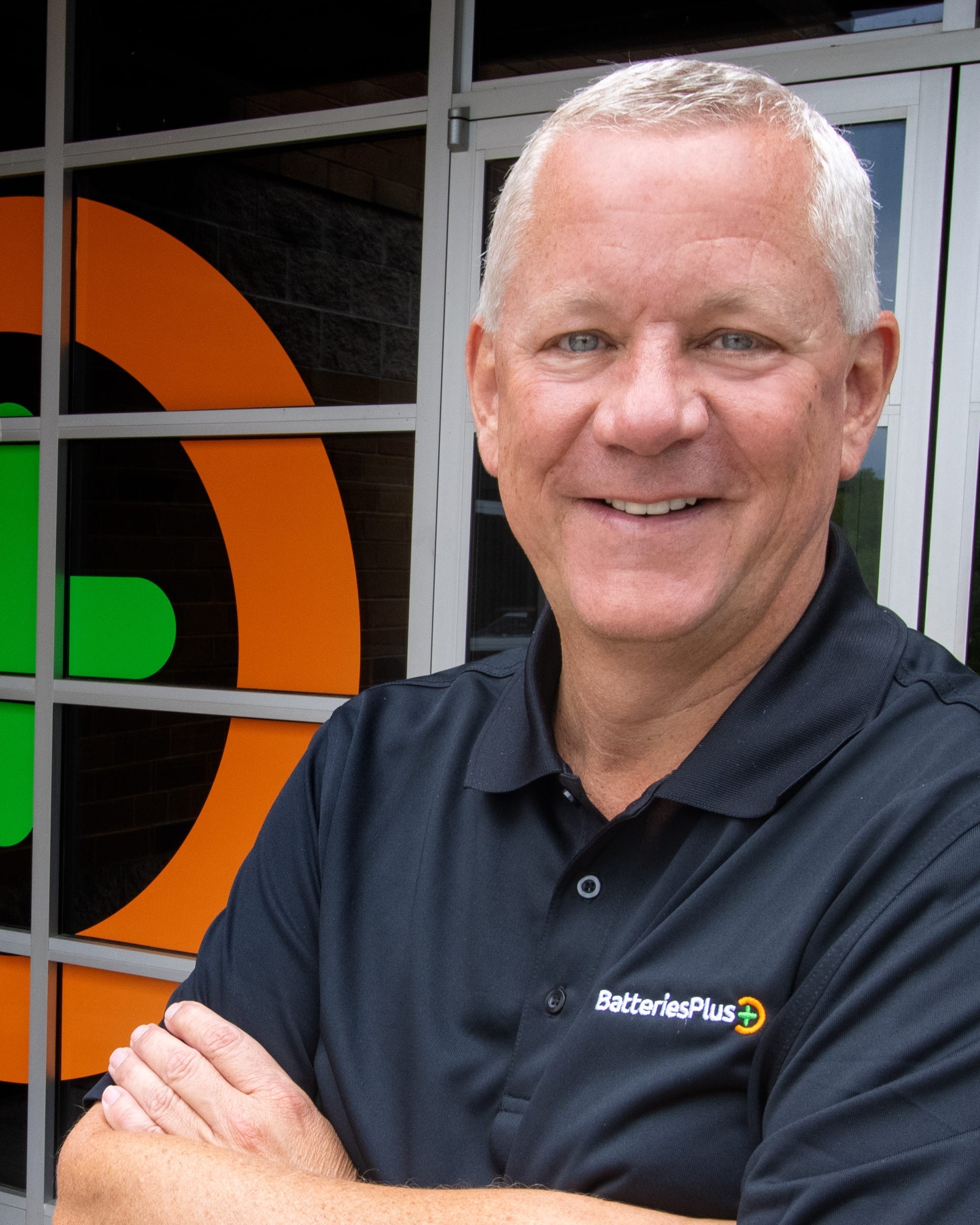 Batteries Plus CEO Scott Williams stands wearing a black polo shirt while crossing his arms while standing in front of a Batteries Plus store