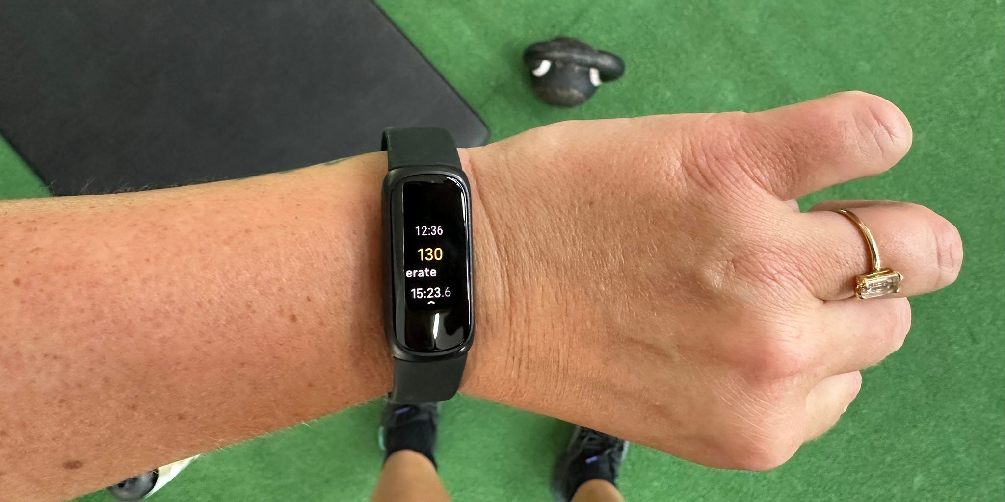 A person wearing a black fitness tracker checks their stats during a workout on green gym flooring, with a black mat and kettlebell visible in the background.