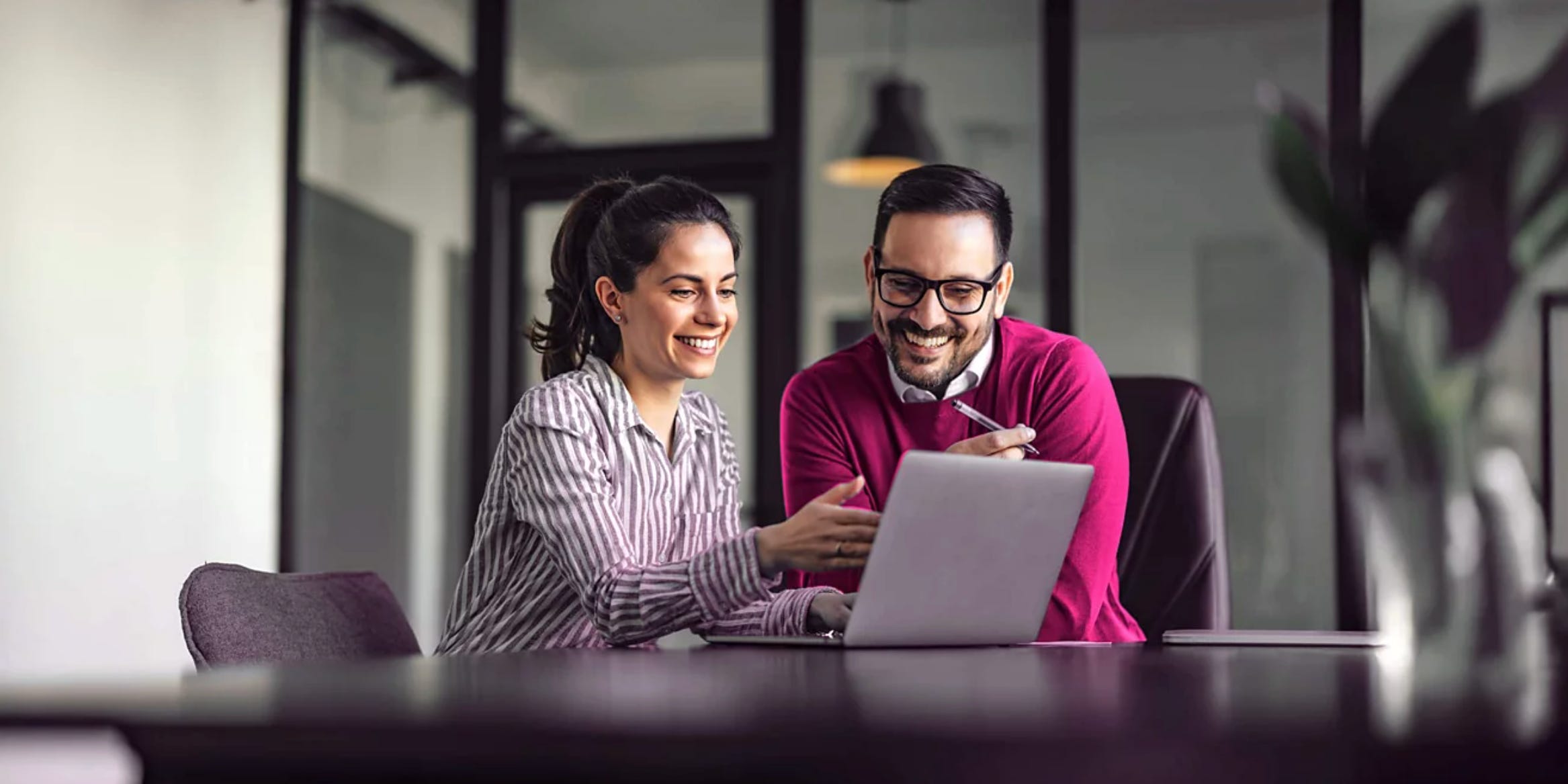 Two people smiling while looking at a laptop together in a modern office setting, using T-Mobile business internet services.