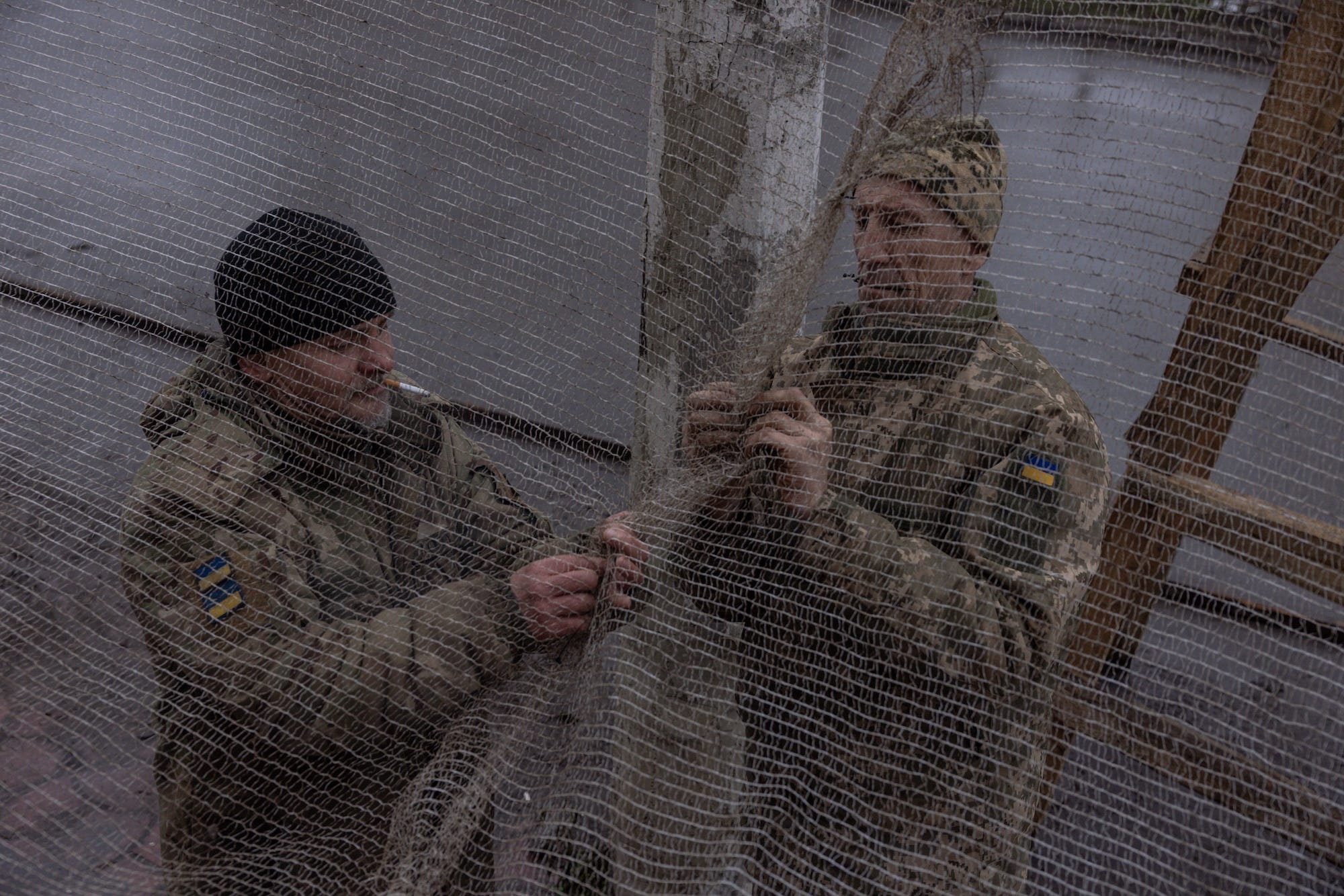 Two men in camoflage outfits and beanies attatch pale-colored netting to a pole