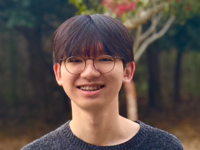 Smiling at the camera, a young person with round glasses and braces stands outdoors against a blurred backdrop of trees.