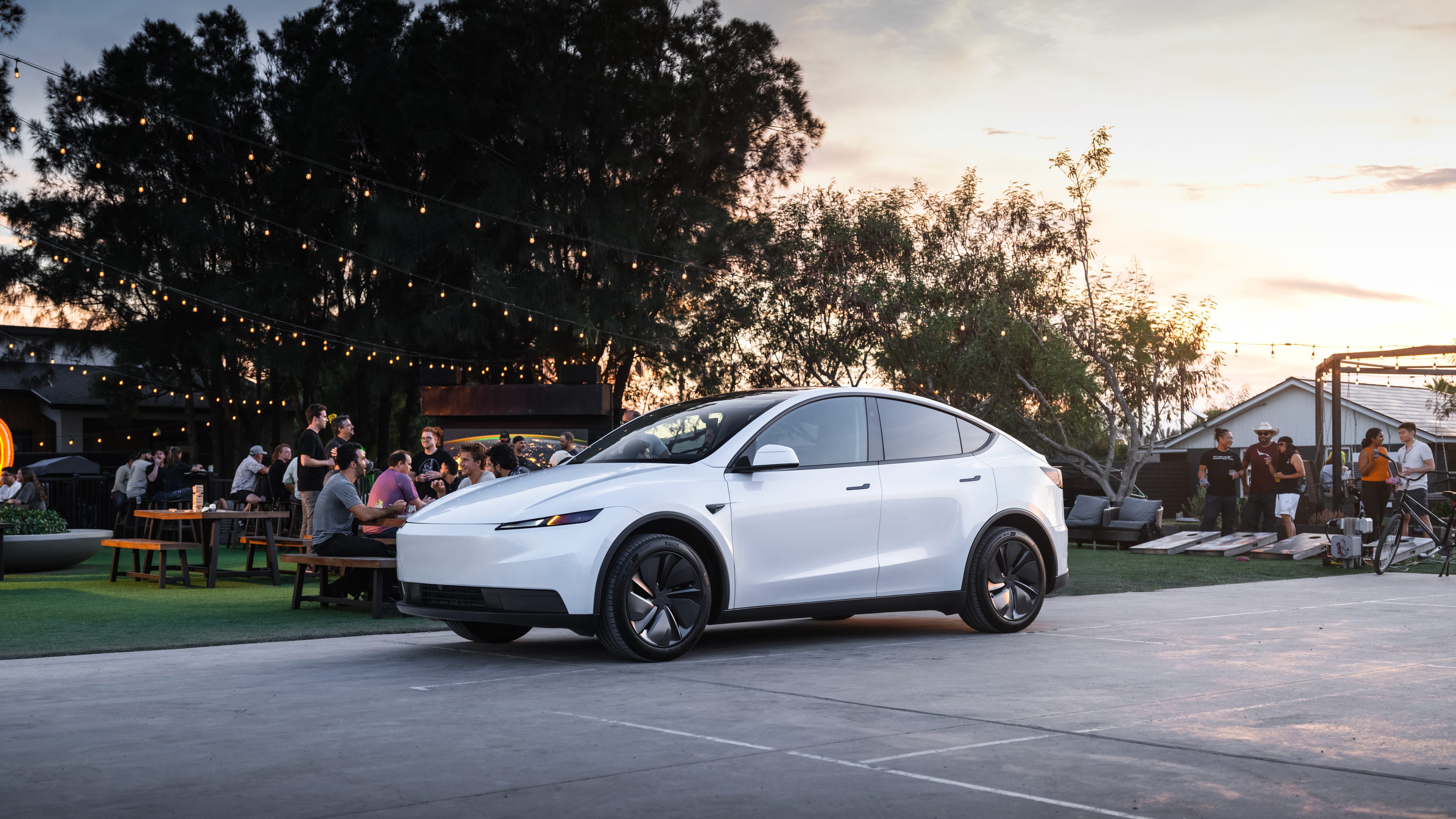 A white Tesla Model Y Standard parked on a gray slab of cement. Behind the car, groups of people are partying with picnic benches and cornhole boards.