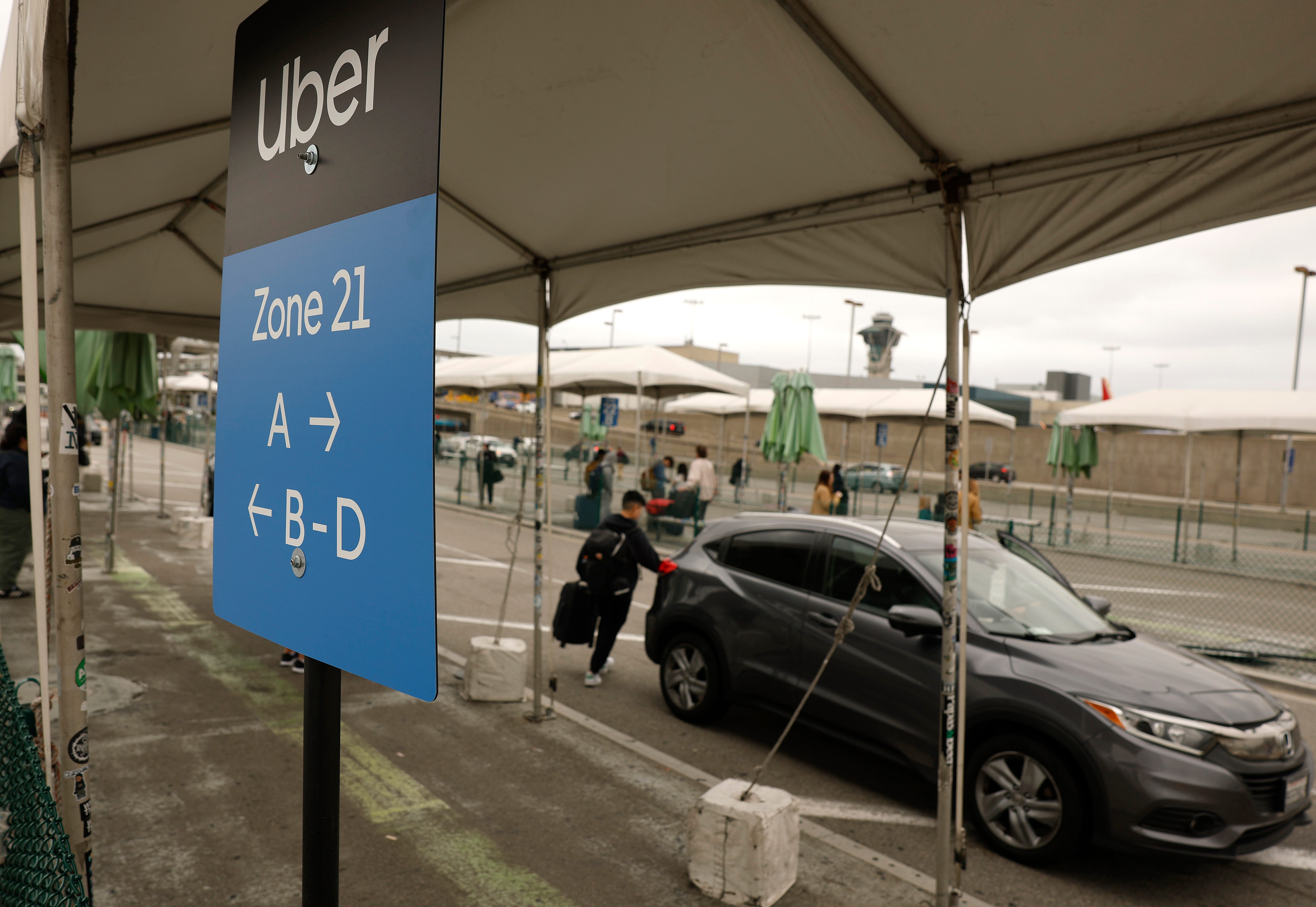 A sign reading "Uber" and pointing passengers toward different pick-up zones labeled by letters stands under a tent as a Honda SUV sits in the background and a passenger with a roller bag walks toward it.